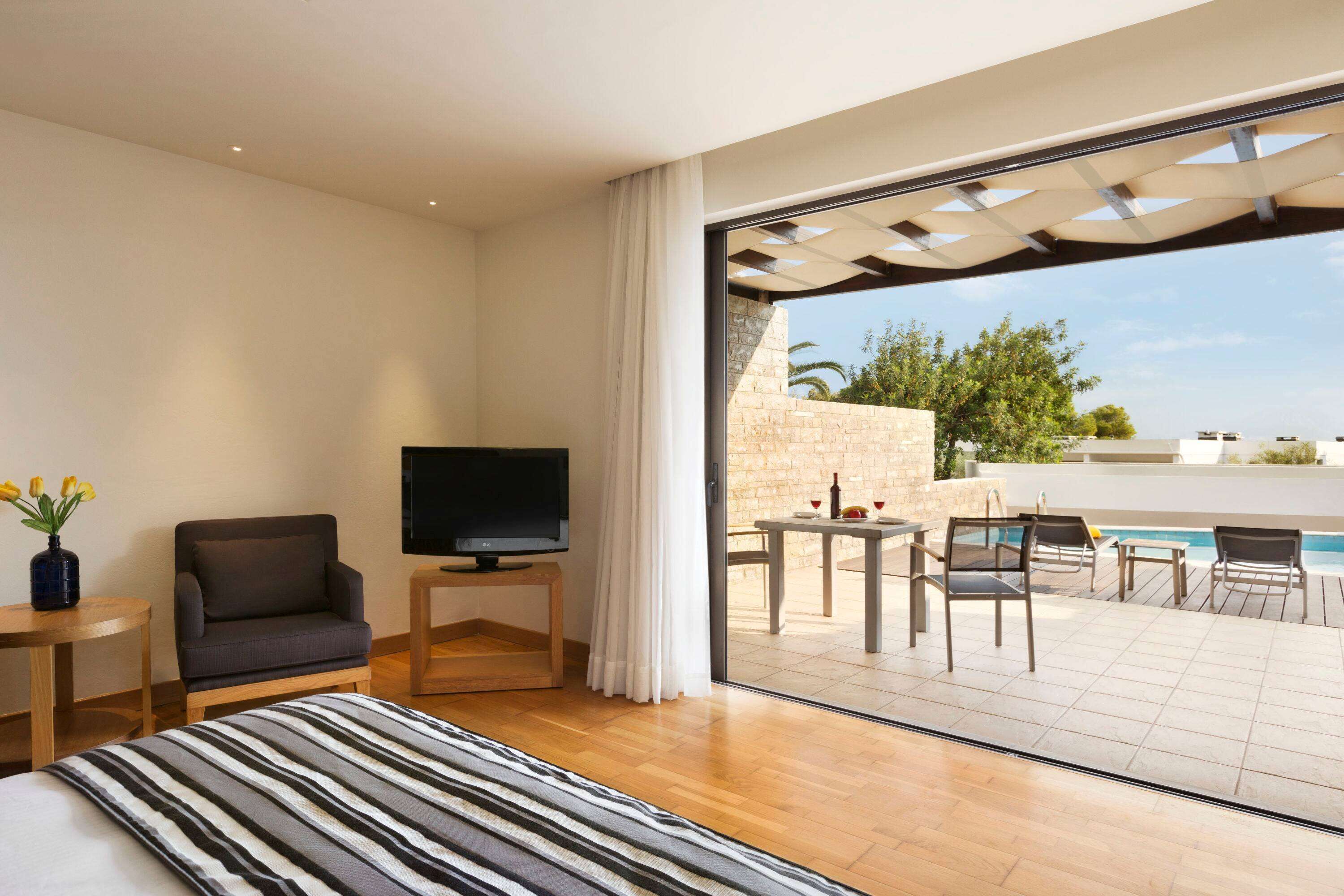 A hotel room with a bed, a TV, a chair, a small table, and a view of an outdoor pool area.