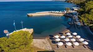 A coastal resort with a clear blue sea, a rocky beach, and a row of sun loungers and umbrellas. - Wyndham Loutraki Poseidon Resort (Loutraki-Agioi Theodoroi)