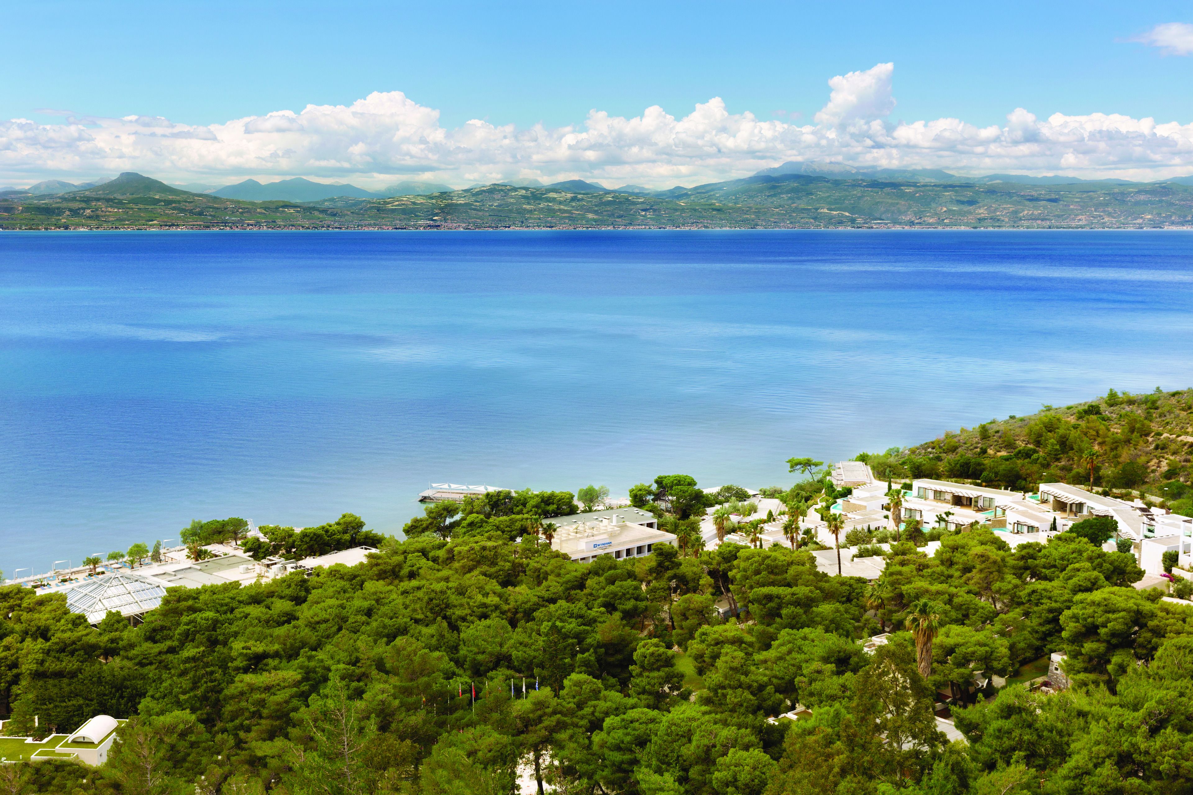 A coastal area with a clear view of the sea, a cluster of buildings surrounded by dense greenery, and distant mountains under a partly cloudy sky.
