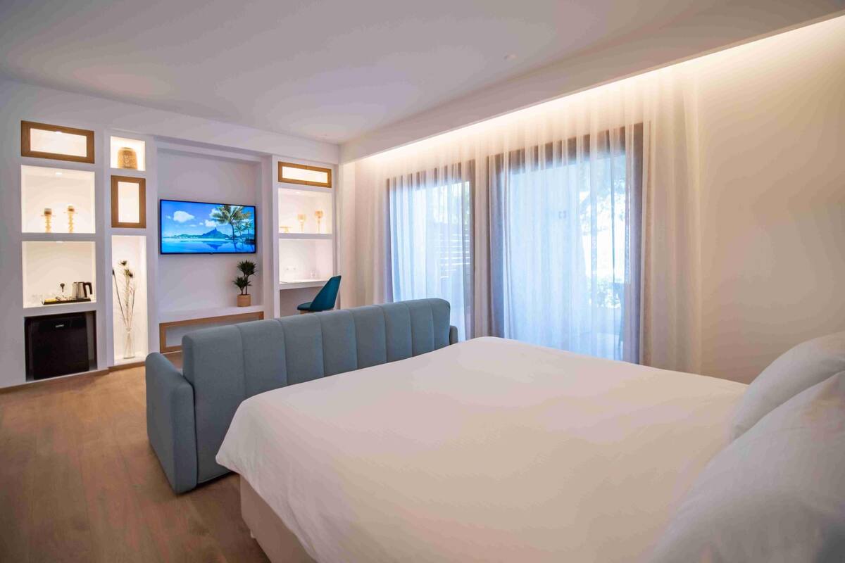 A modern hotel room with a large bed, a flat-screen TV mounted on the wall, a seating area with a blue sofa, and a wooden cabinet with decorative items.