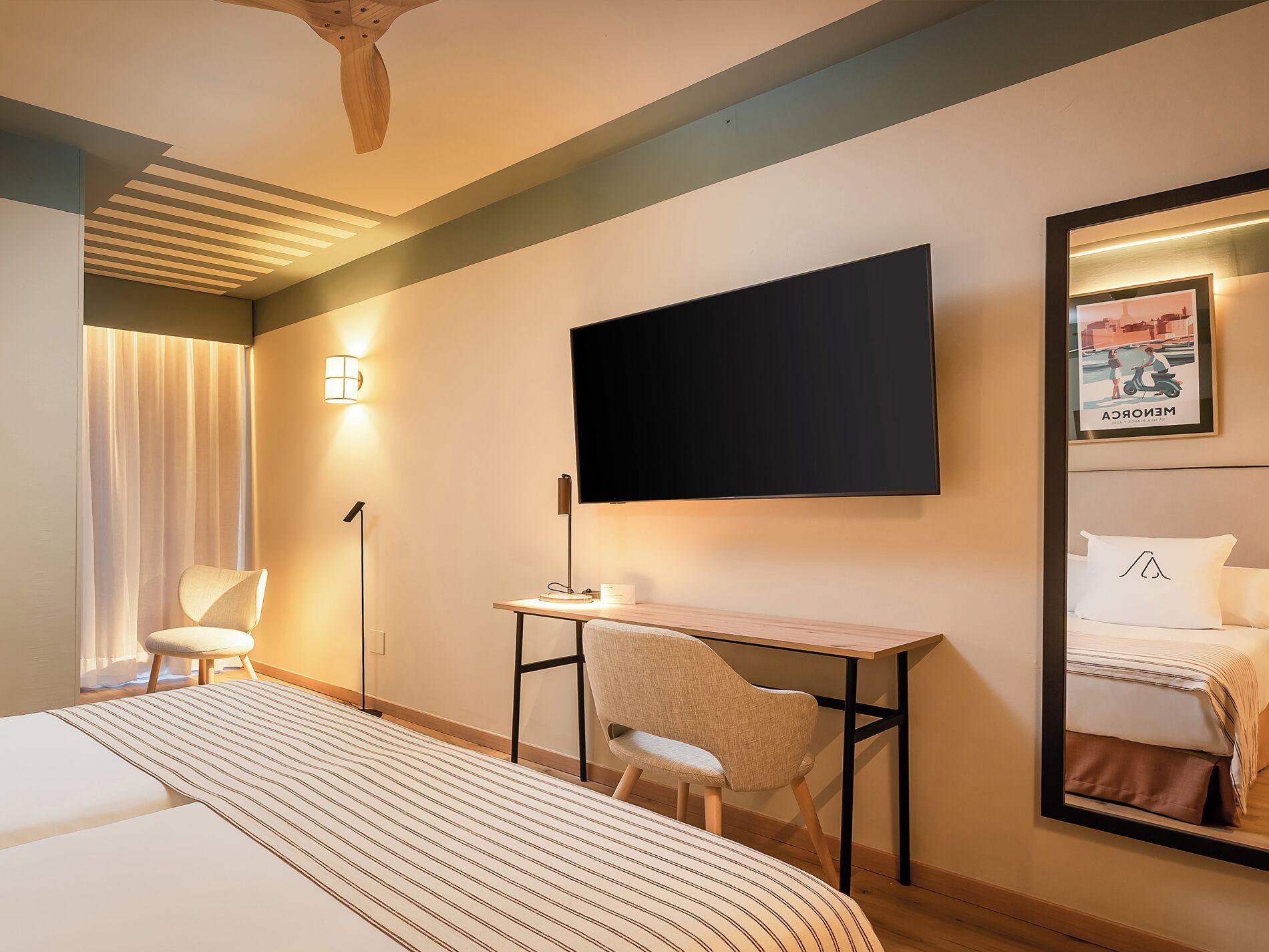 Habitación de hotel con cama, escritorio, silla, televisor de pantalla plana, lámpara, espejo y un cartel en la pared.