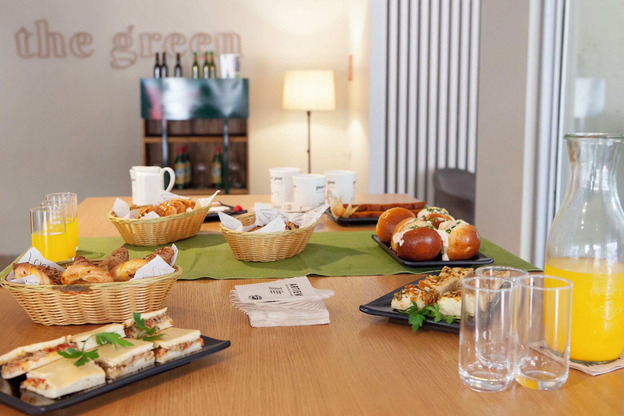 A breakfast buffet with various pastries, sandwiches, and a pitcher of orange juice on a wooden table.