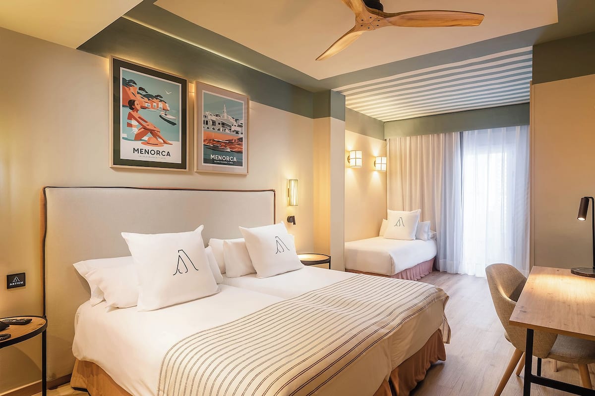 A hotel room with a large bed, a smaller bed, a desk, and a ceiling fan. There are two framed pictures on the wall and a window with curtains.