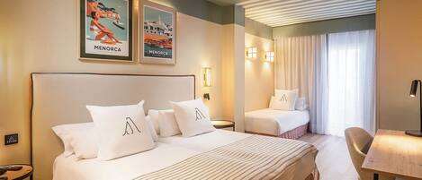 Habitación de hotel con una cama grande, una cama más pequeña, un escritorio y un ventilador de techo. Hay dos cuadros en la pared y una ventana con cortinas.
