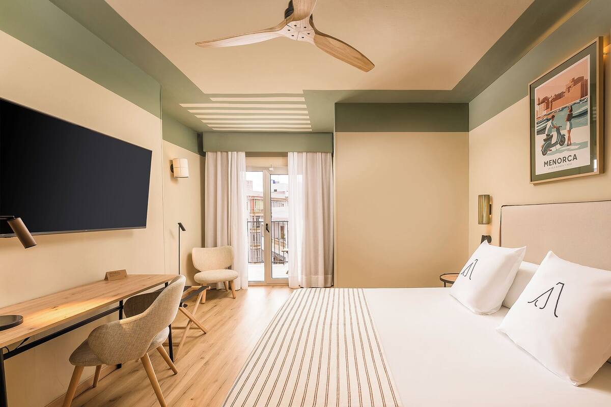 A hotel room with a bed, a desk, a chair, a ceiling fan, and a view of the outside.