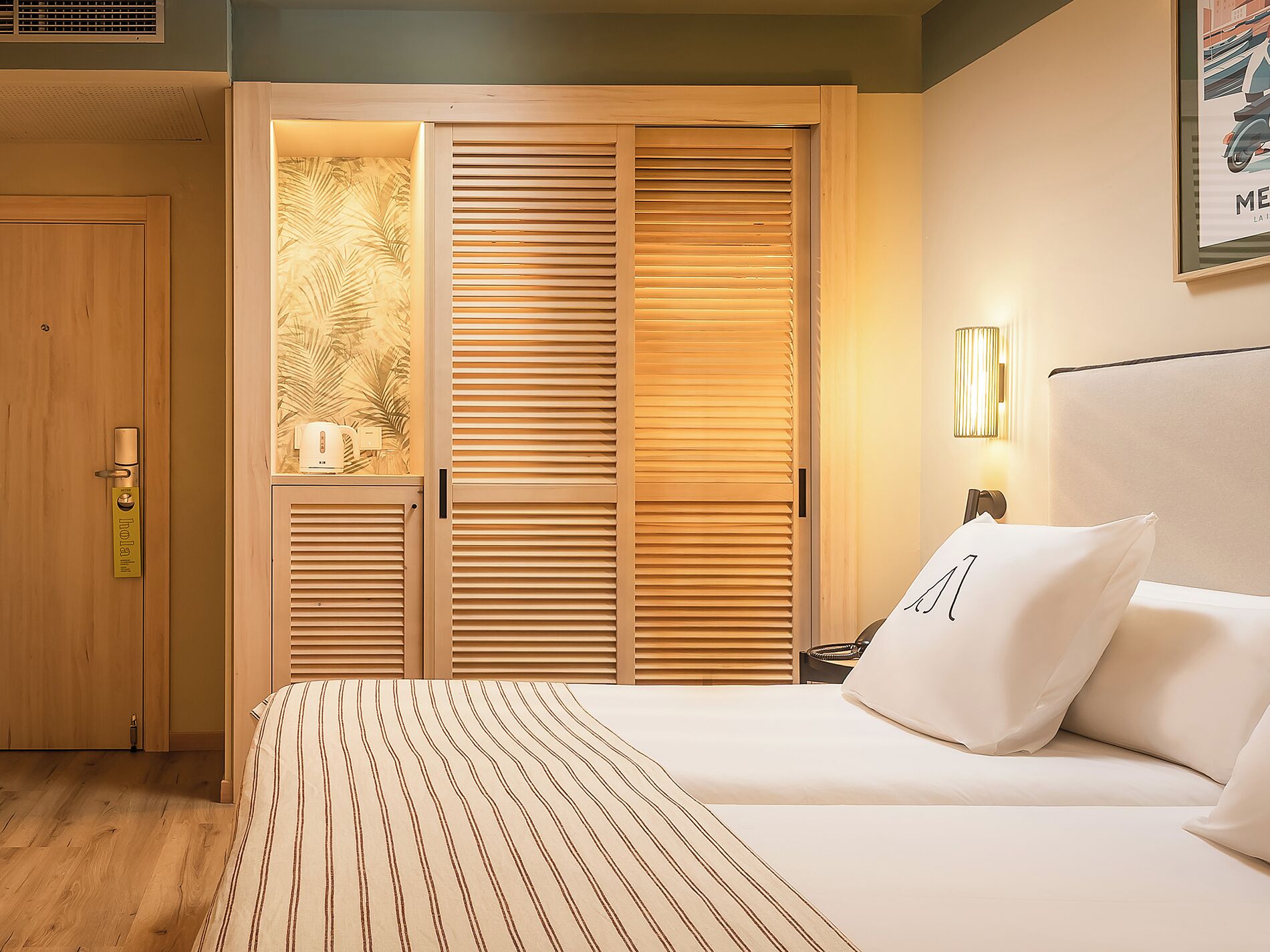 A hotel room with a bed, a nightstand, a lamp, and a wooden wardrobe with a palm leaf design.