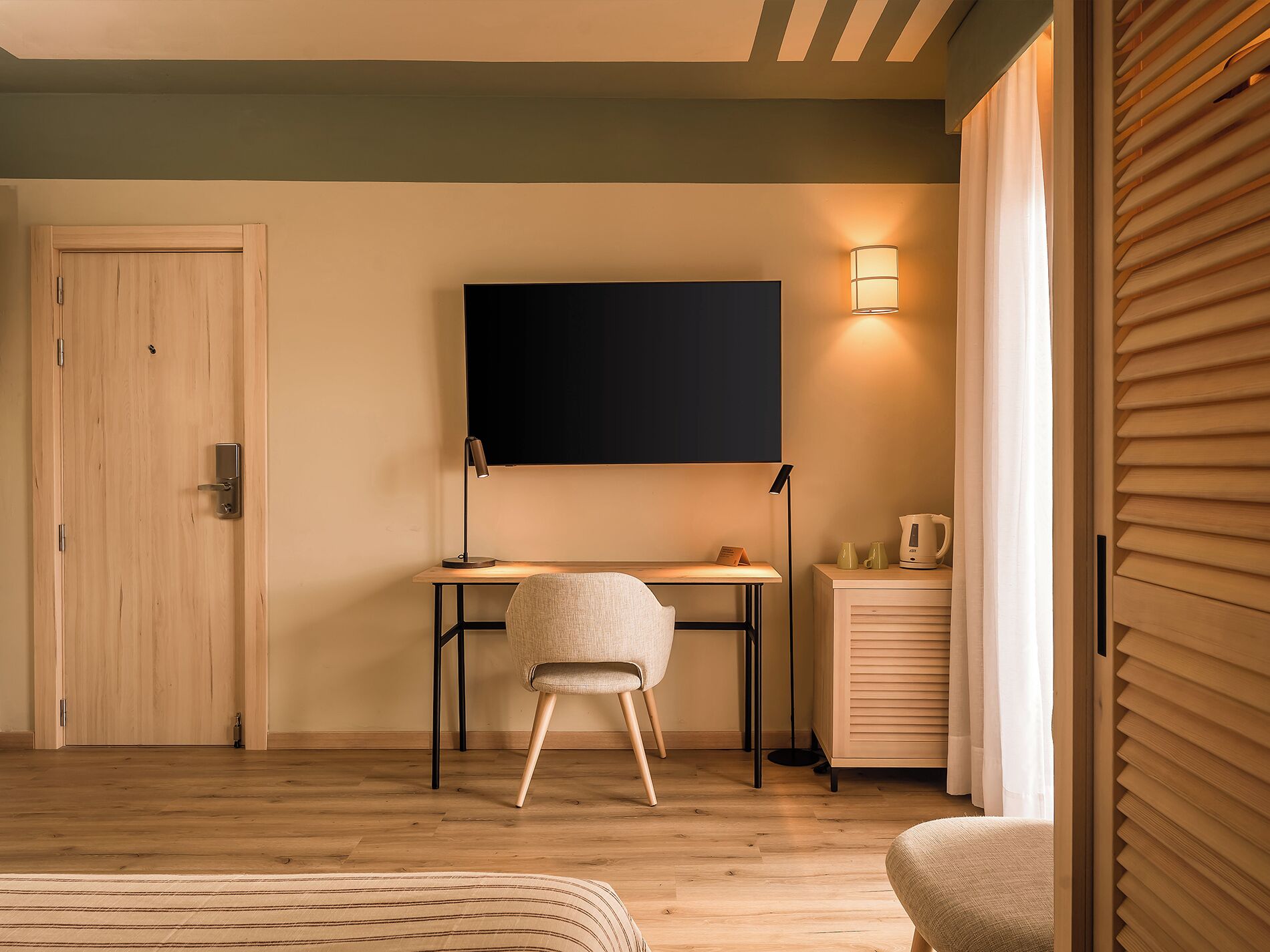 A room with a wooden desk, a chair, a flat-screen TV, a lamp, and a bedside table with a kettle.