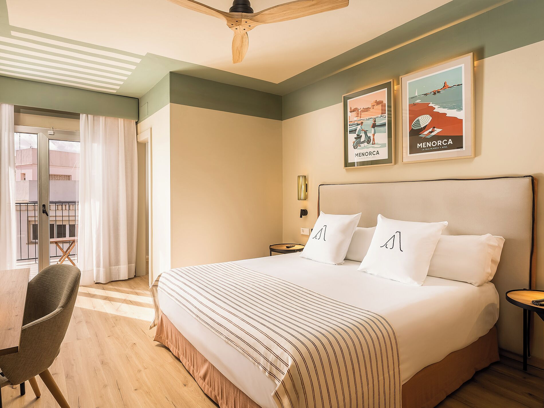 A hotel room with a large bed, a chair, a small table, and a ceiling fan. There are two framed pictures on the wall.