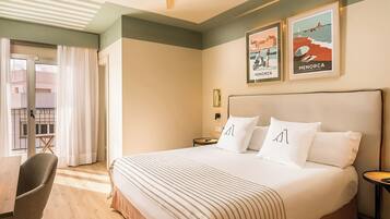 A hotel room with a large bed, a chair, a small table, and a ceiling fan. There are two framed pictures on the wall.
