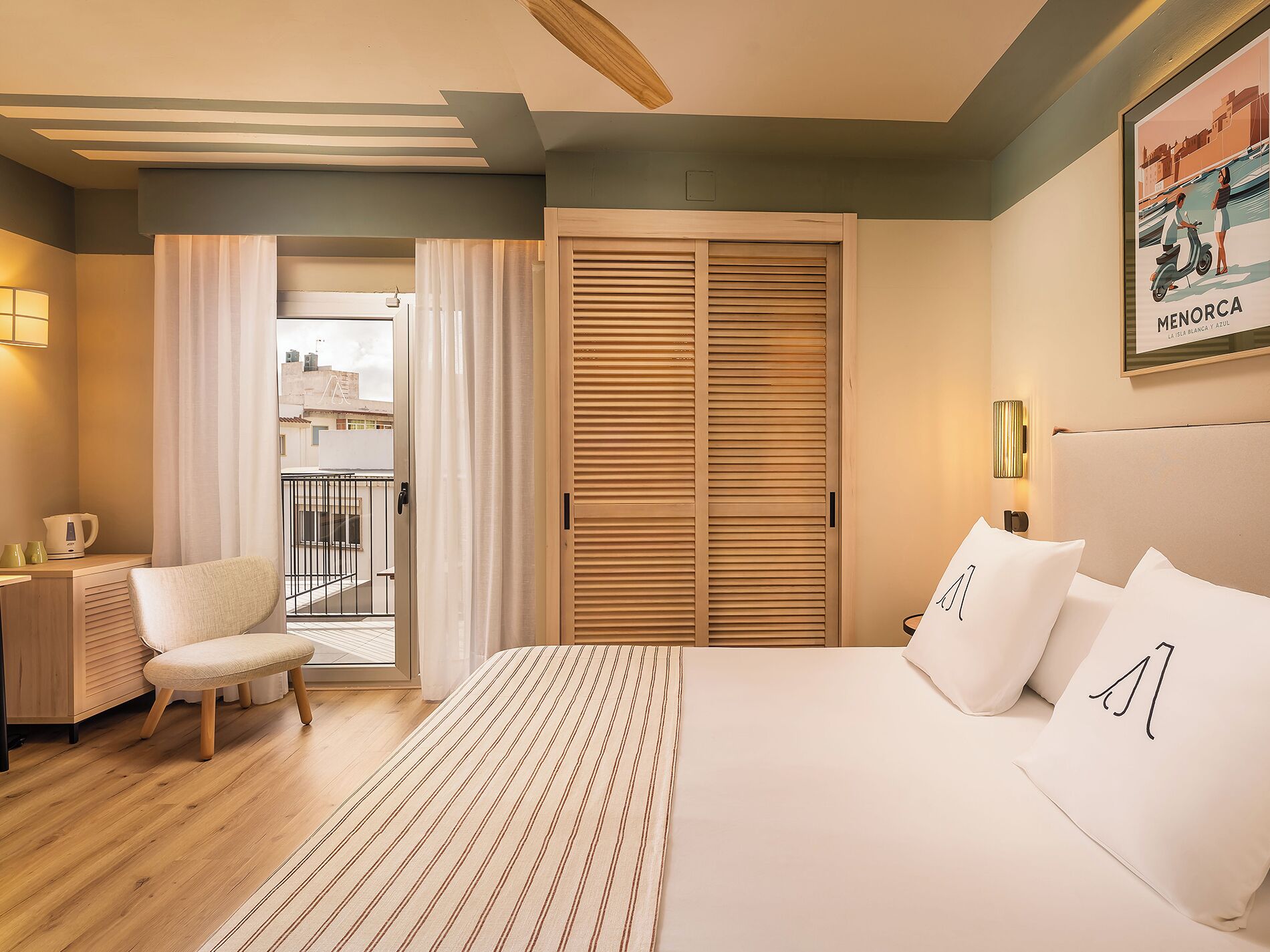 Habitación de hotel con cama, una silla, escritorio con lámpara, ventana con cortinas y balcón.