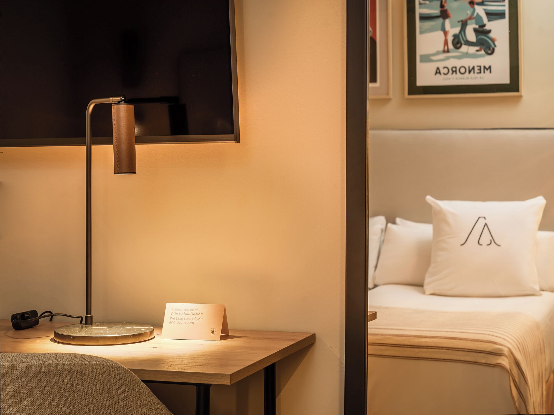 A hotel room with a desk, a lamp, a bed with white bedding, and a framed picture on the wall.