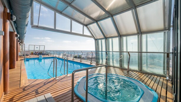 Un espace piscine intérieur d’un navire de croisière, doté d’un jacuzzi, d’une terrasse en bois et de grandes baies vitrées offrant une vue sur la mer.