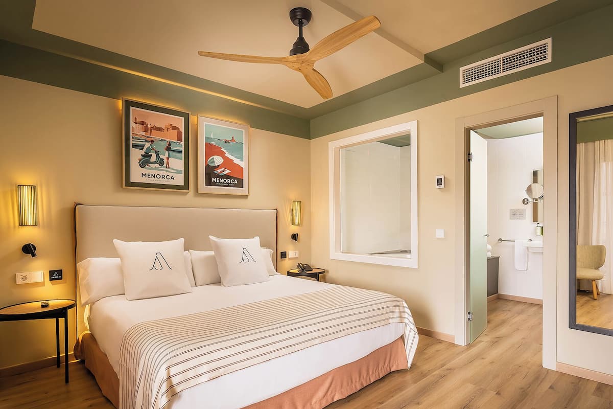 A hotel room with a large bed, a ceiling fan, framed pictures on the wall, and a view into the bathroom.