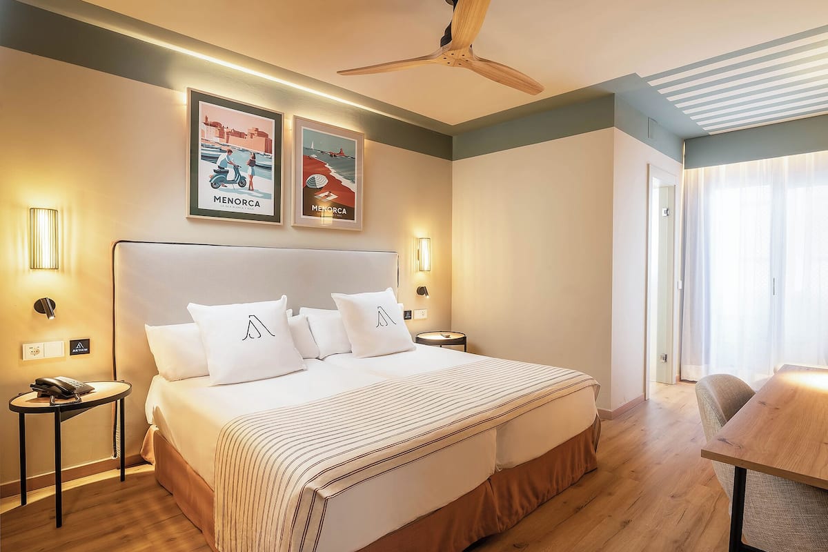 A hotel room with a large bed, a ceiling fan, framed pictures on the wall, and a wooden table.