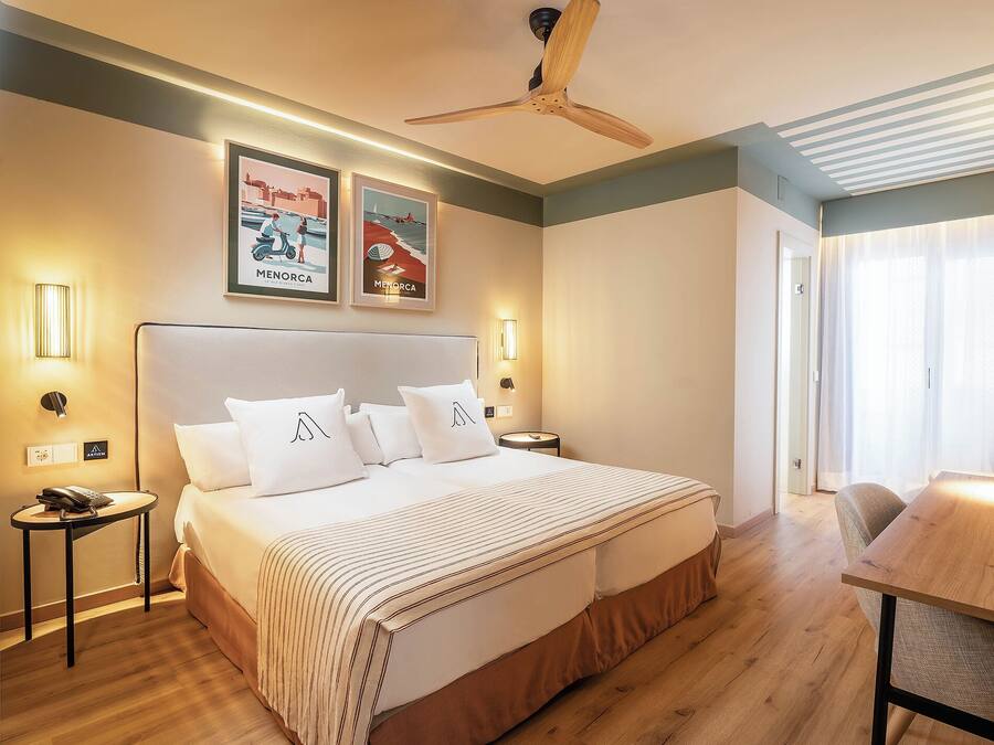 A hotel room with a large bed, a ceiling fan, framed pictures on the wall, and a wooden table.