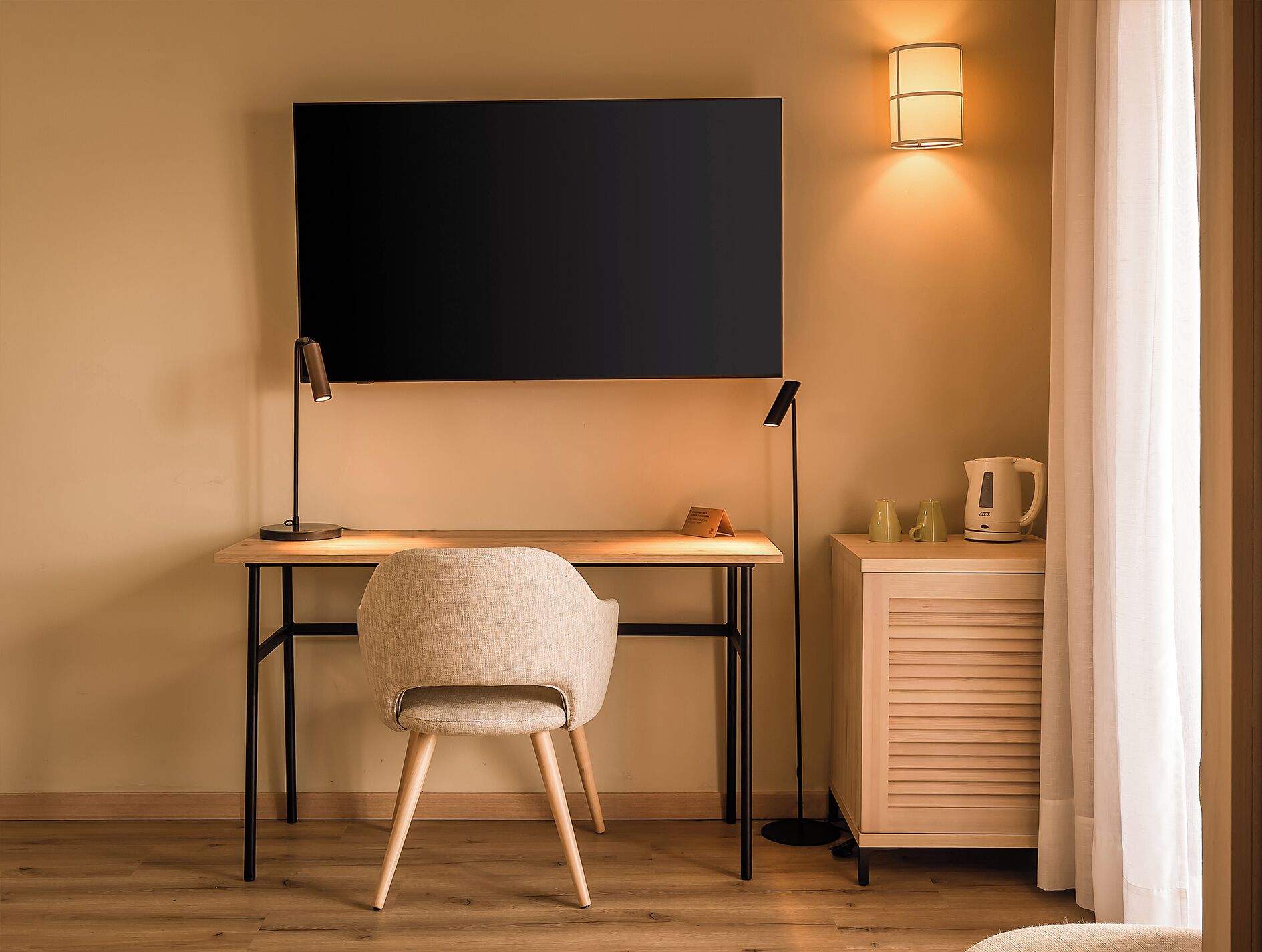 Una habitación moderna con un escritorio de madera, una silla, un televisor de pantalla plana montado en la pared, una lámpara y un mueble con una tetera.