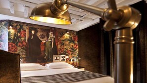 Una cama con una manta a rayas, una pared con papel tapiz floral y una lámpara de bronce.