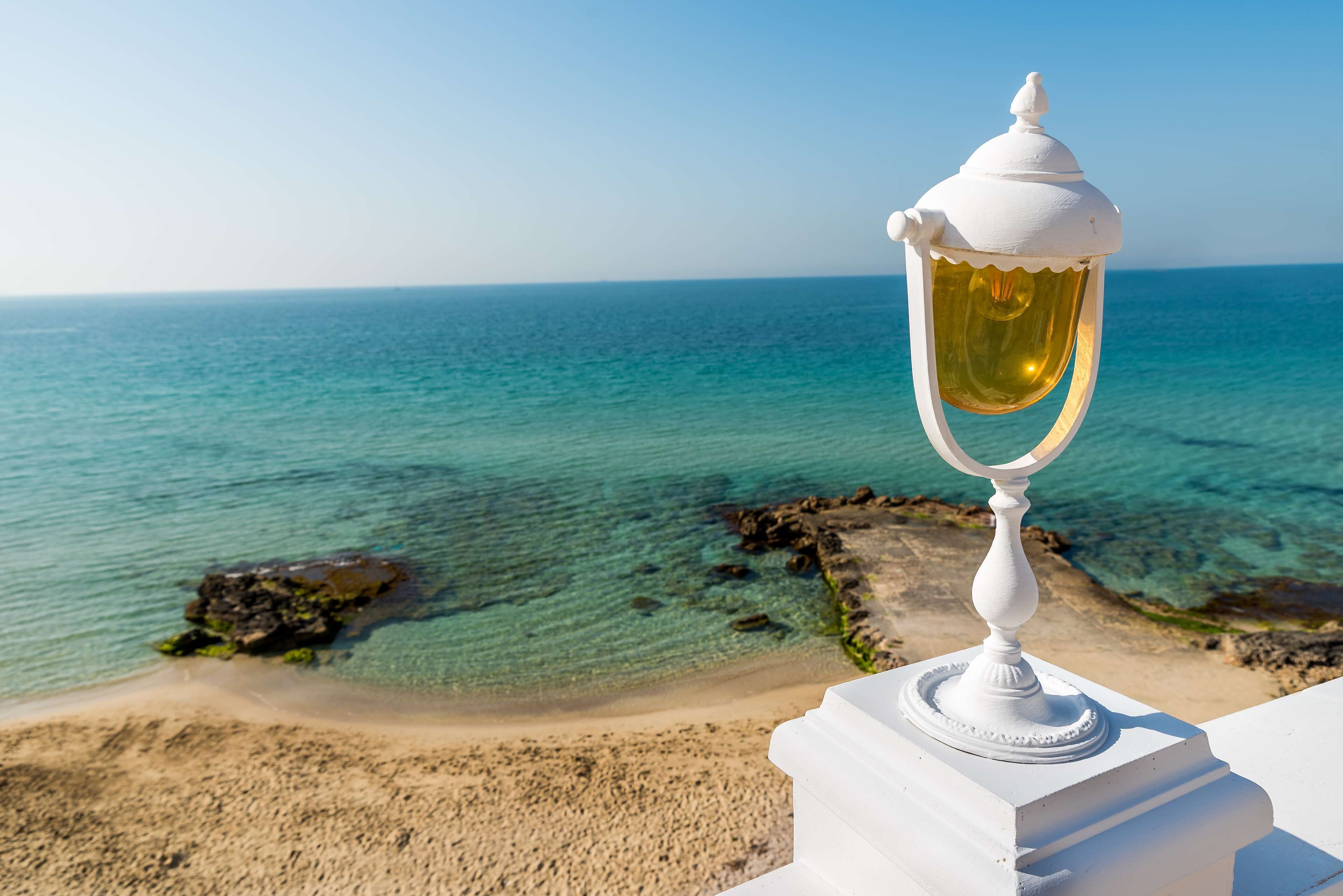 Una lámpara blanca sobre un pedestal blanco, con vista a una playa de aguas turquesas cristalinas y afloramientos rocosos.