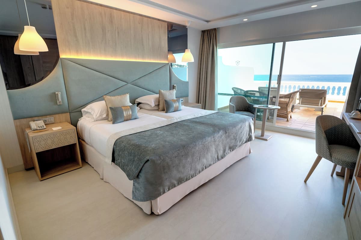 A modern hotel room with a large bed, a desk, and a balcony with a view of the sea.