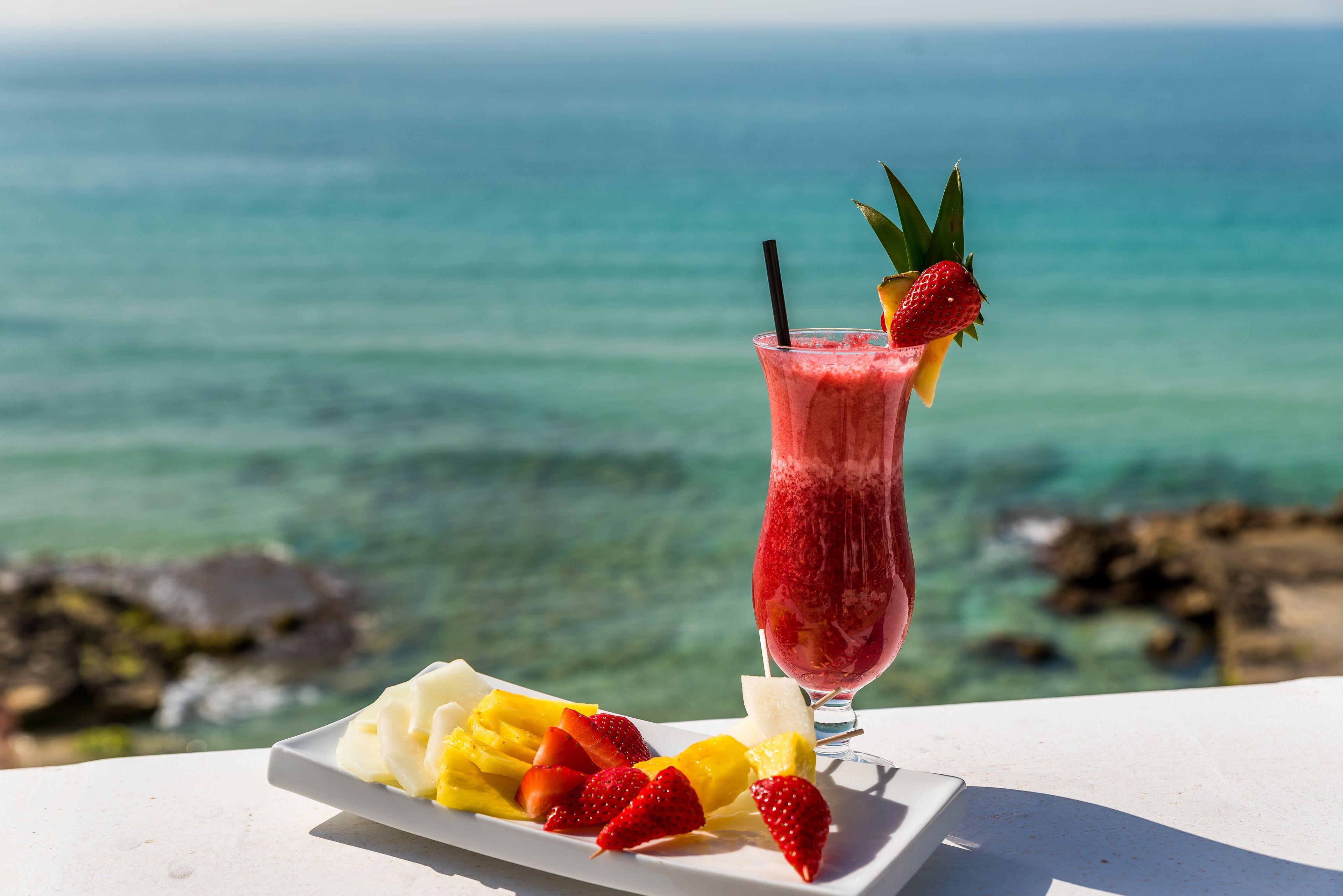 Ein Glas roter Fruchtsaft mit Trinkhalm und Erdbeere als Dekoration, ein Teller mit Früchten, darunter Erdbeeren und Mango, sowie Blick aufs Meer.