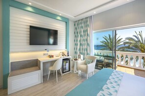 Una habitación de hotel moderna con un televisor grande, una kitchenette pequeña, un balcón con vistas al océano y una cama con colcha de diseño azul y blanco.