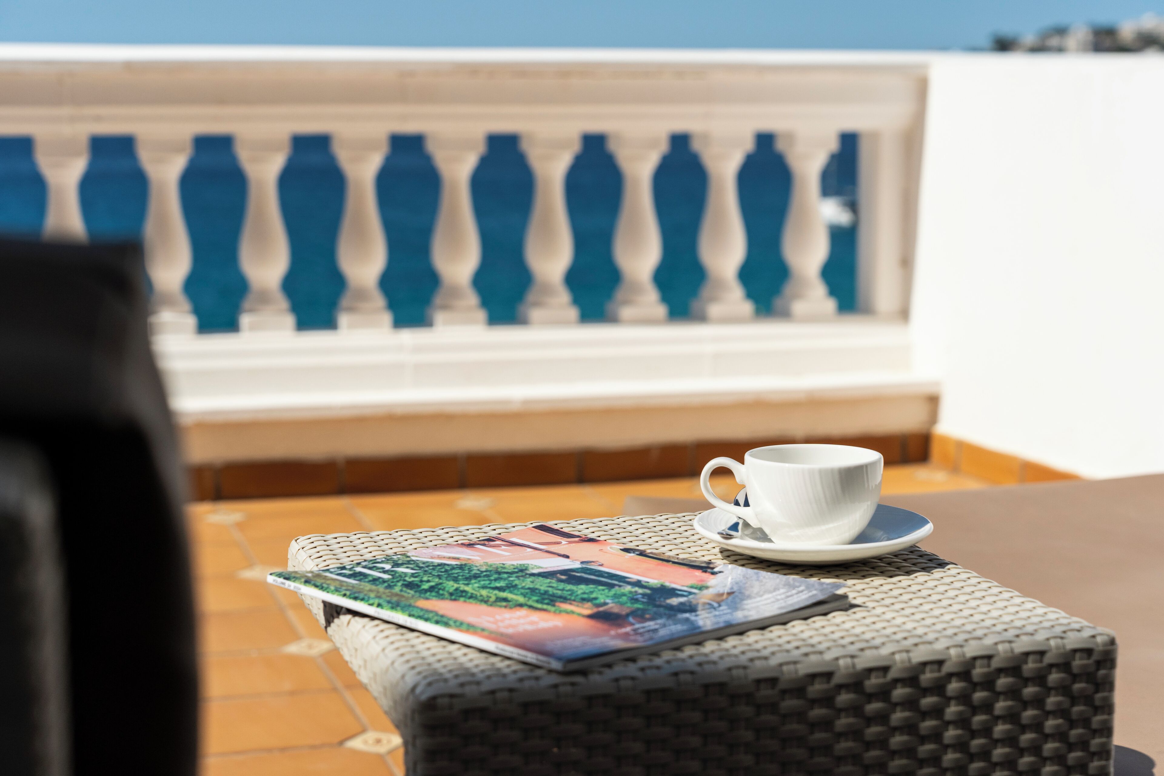 A white cup on a saucer on a woven table, with a magazine and a cushion.