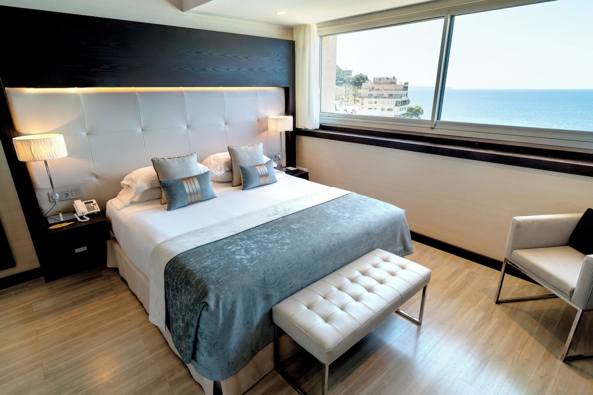 A modern hotel room with a large bed, a bench, a chair, a bedside table with a lamp, and a view of the sea.