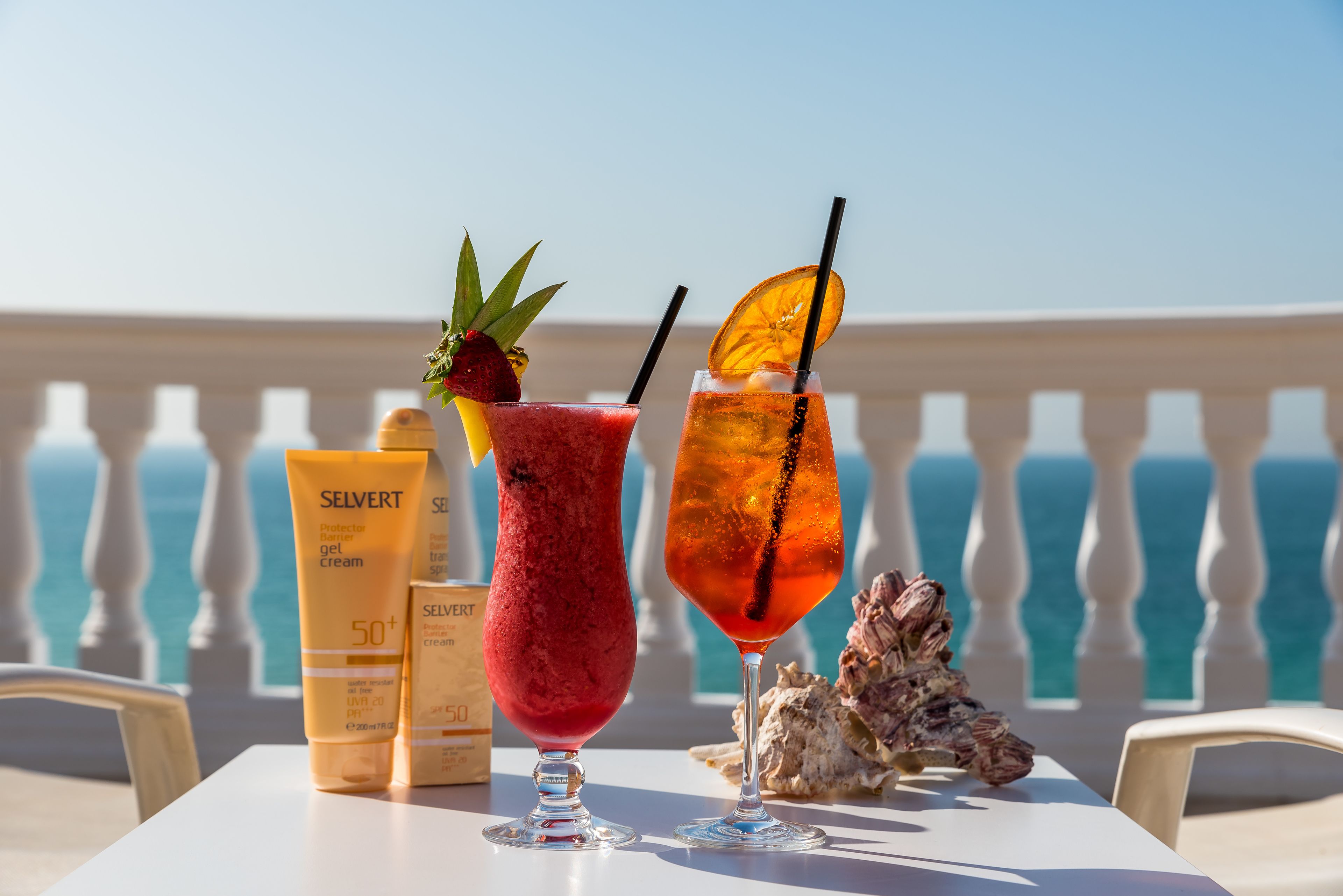 Un tavolo con due bevande, crema solare e una conchiglia su un balcone che si affaccia sul mare.