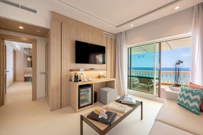 A modern hotel room with a flat-screen TV mounted on a wooden panel wall, a small refrigerator, a seating area with a sofa and a coffee table, and a balcony with a view of the sea.