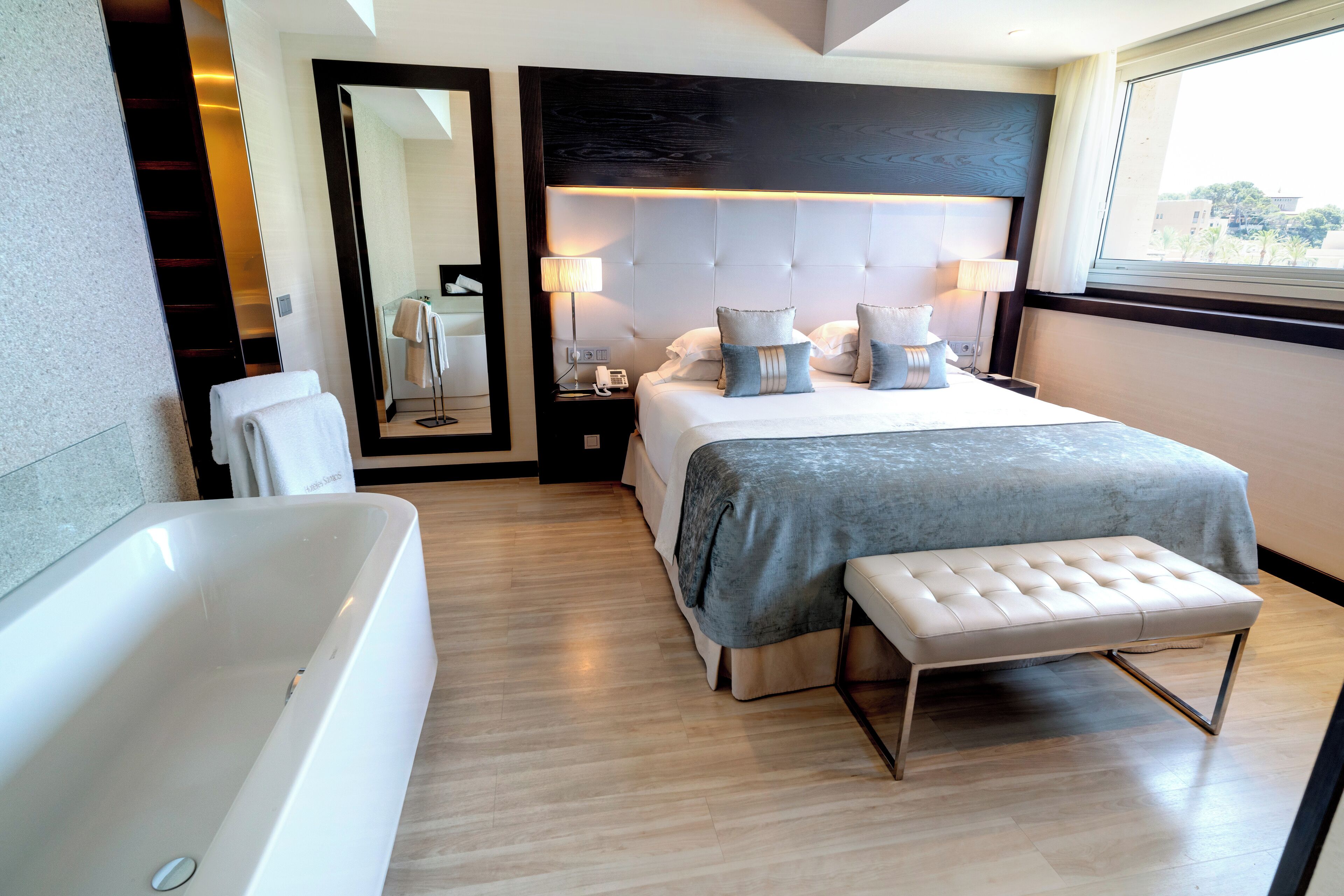 Una habitación de hotel moderna con una cama grande, un banco para sentarse, una bañera y un espejo amplio.