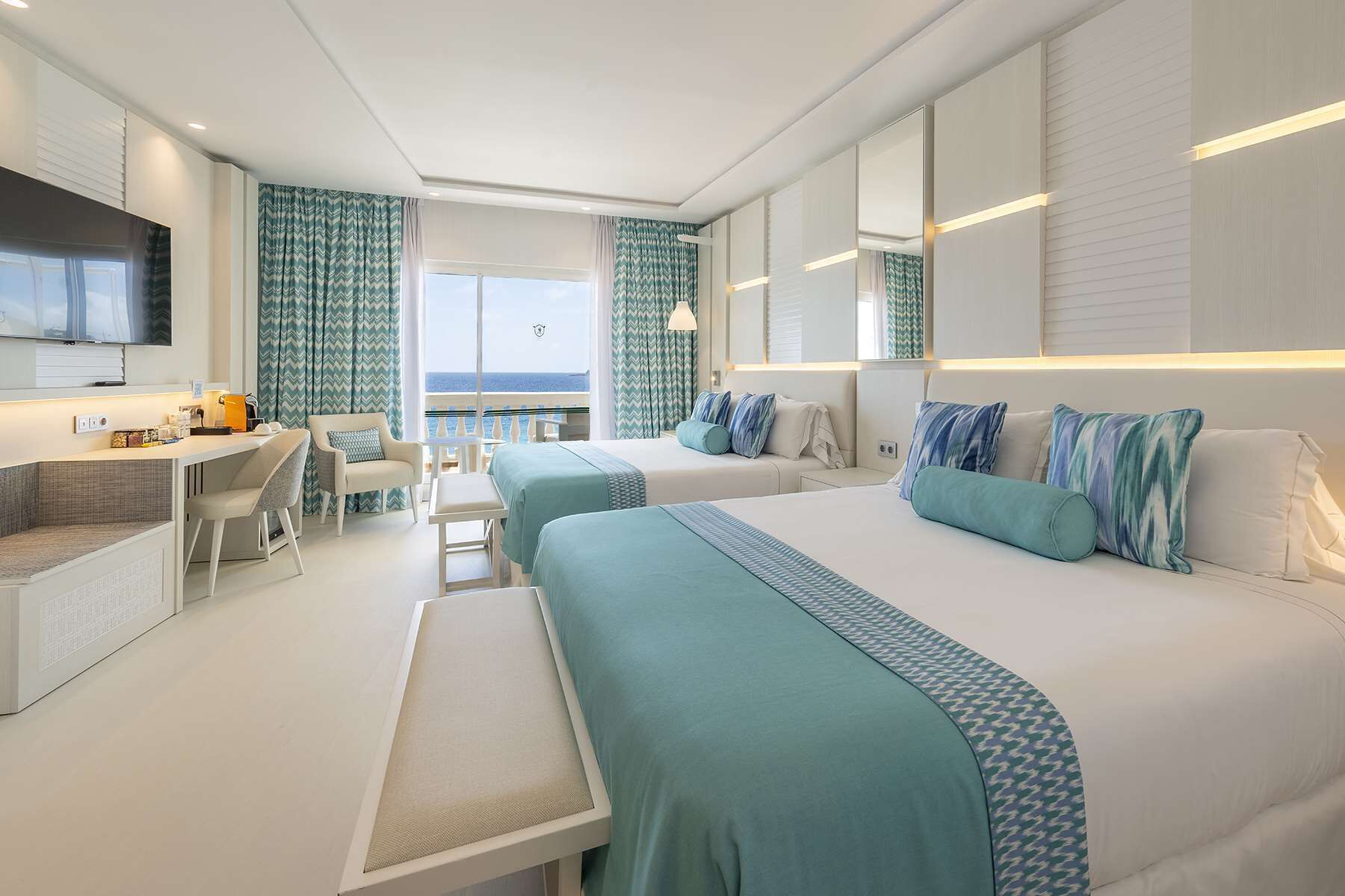 A modern hotel room with two beds, a desk, and a balcony with a view.