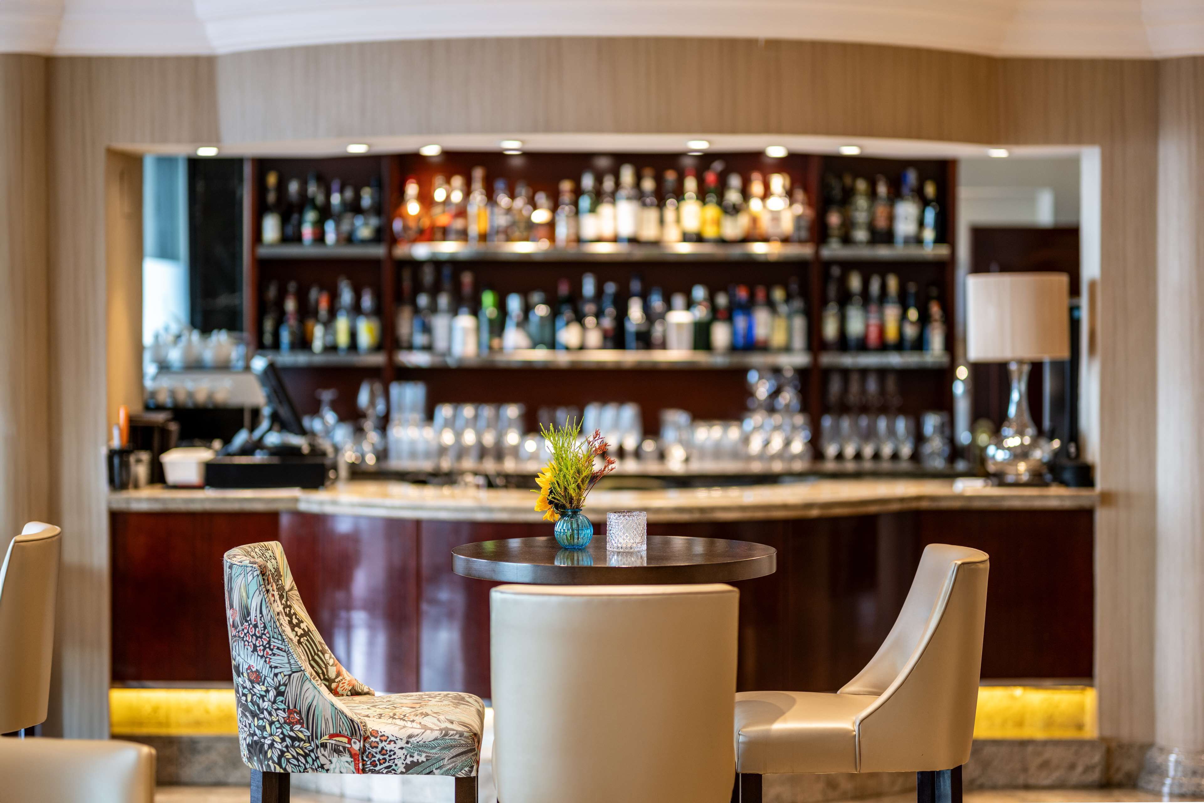 Una zona de bar con una mesa redonda y dos sillas, un jarrón con flores y una estantería bien surtida al fondo.
