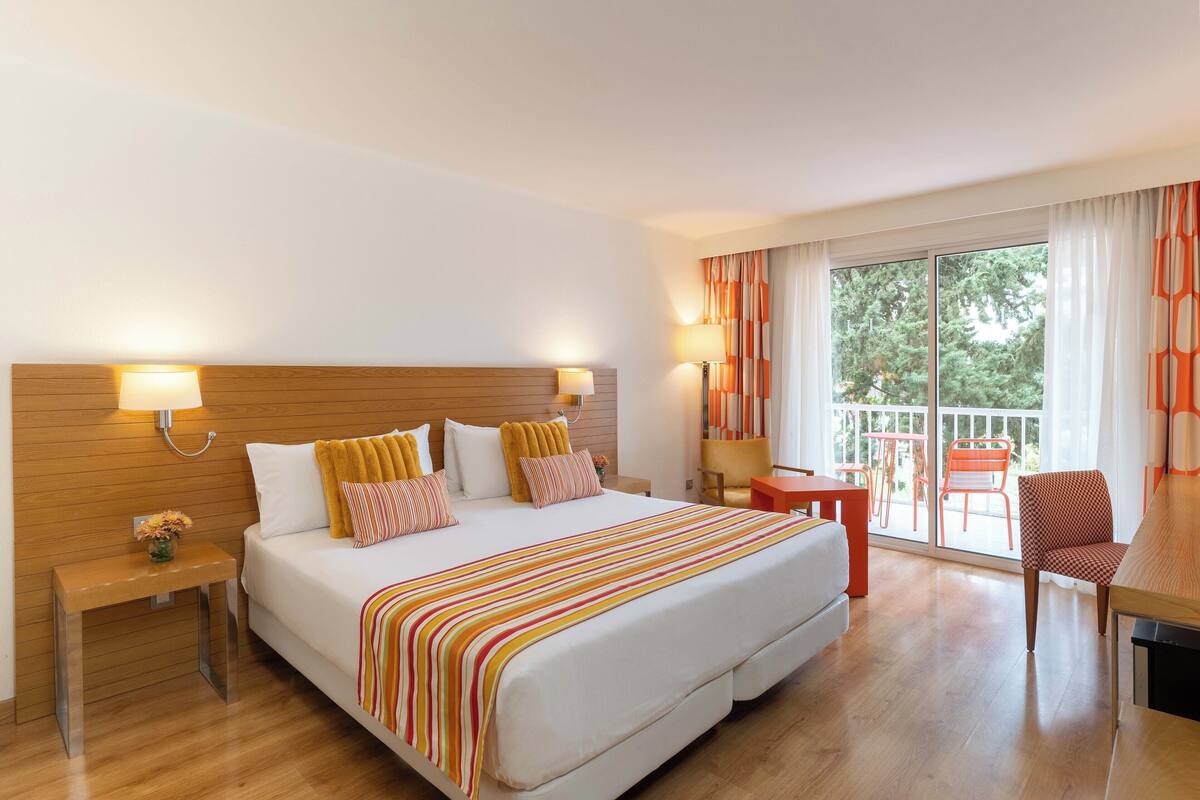 A hotel room with a large bed, wooden headboard, two bedside tables, a small table, and a chair. There is a balcony with orange chairs and a table.