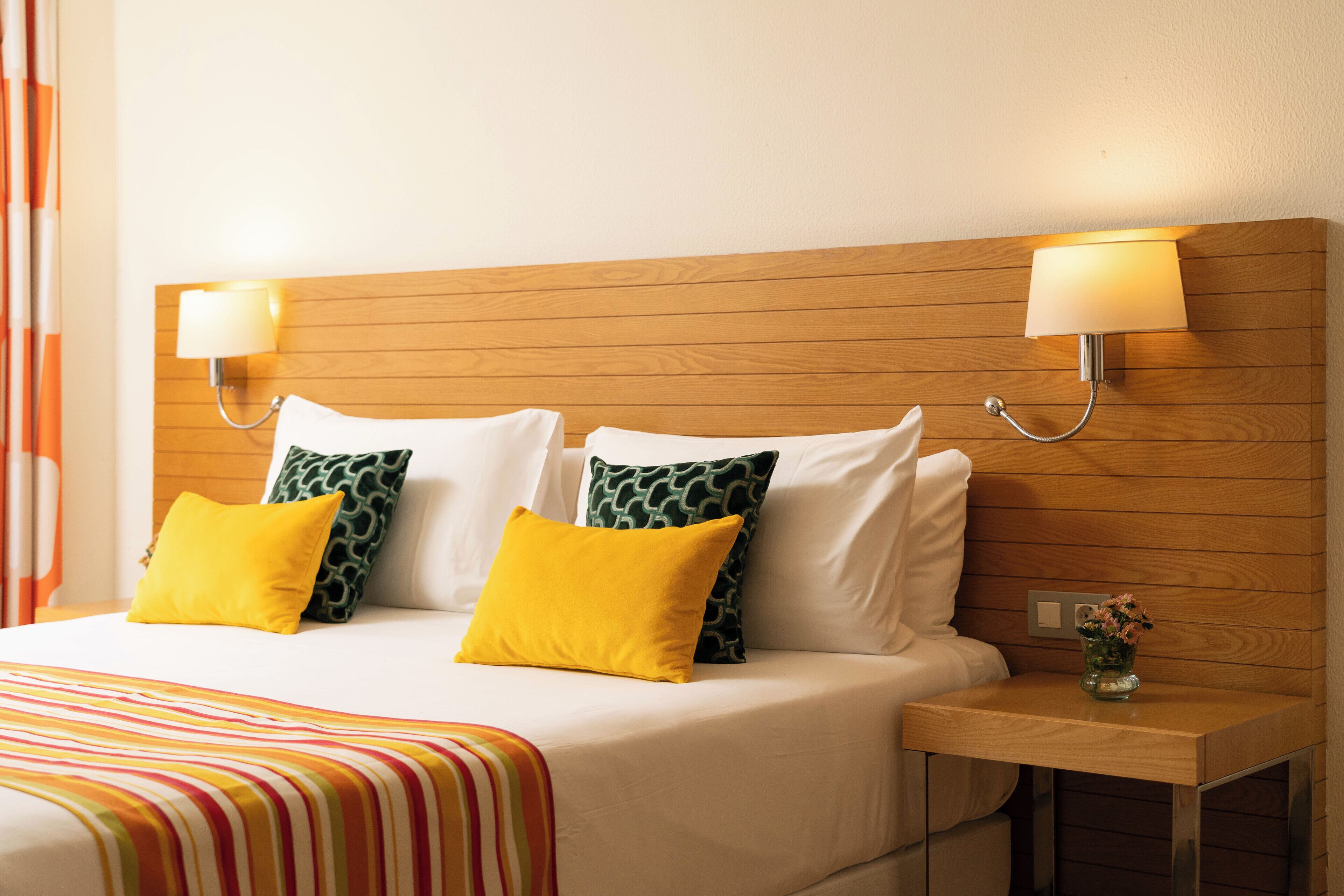 Ein Hotelzimmer mit einem hölzernen Kopfteil, zwei Nachttischlampen, einem Bett mit weißen und gelben Kissen, einer gestreiften Decke und einer kleinen Vase mit Blumen auf einem Nachttisch.