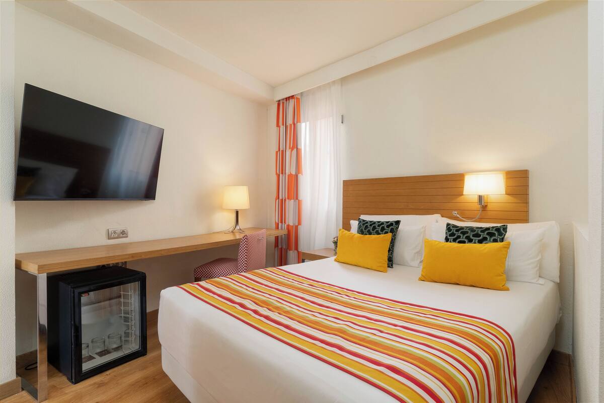 A hotel room with a bed, bedside lamps, a TV, a small refrigerator, and a desk with a chair.