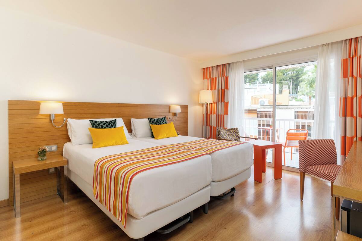 A hotel room with two beds, a wooden headboard, a small table, a chair, and a view of the outside through large windows.