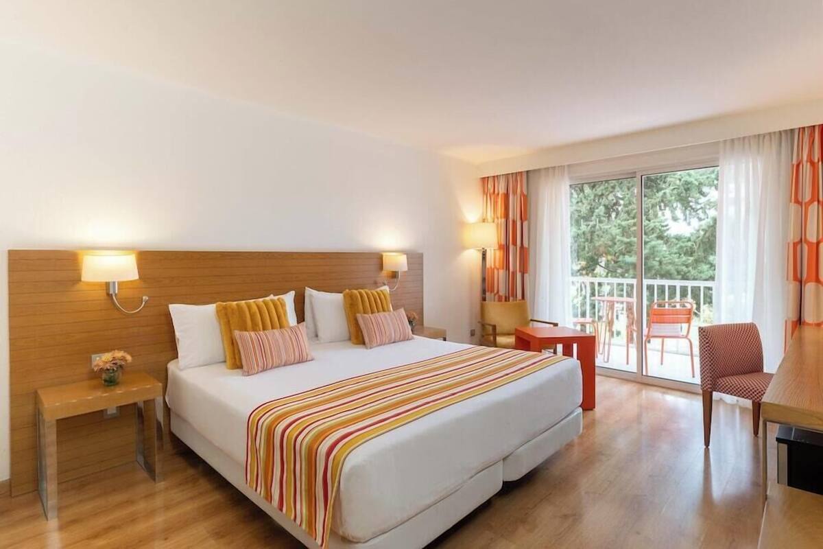 A hotel room with a large bed, bedside lamps, a small table, a chair, and a balcony with a view of trees.