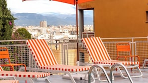 Un área de piscina en la azotea con sillones reclinables a rayas y sombrillas naranjas, con vista a un paisaje urbano.