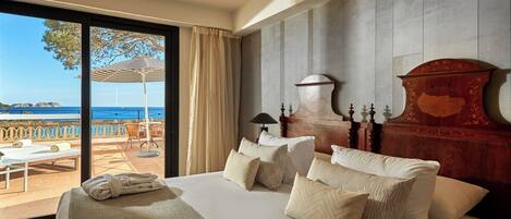 A hotel room with a large bed, a wooden headboard, a view of the sea, and a balcony with outdoor seating.