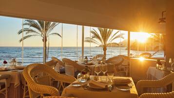 Un comedor con sillas y mesas de mimbre, ventanas amplias con vista al mar y palmeras, y un atardecer al fondo.