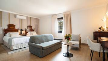 A hotel room with two beds, a sofa, a small round table with a vase of flowers, a desk with a lamp, and a window with curtains.