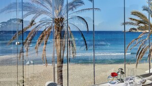 A beach view through a glass window, palm trees, and a table with a glass of water and a candle. - Secrets Mallorca Villamil Resort & Spa - Adults Only (Calvia)