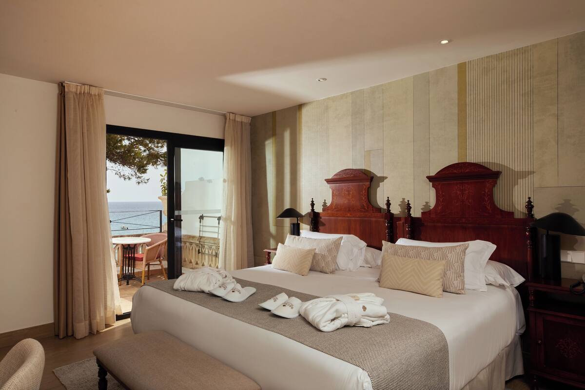 A hotel room with a large bed, a bench, a desk, and a view of the sea.