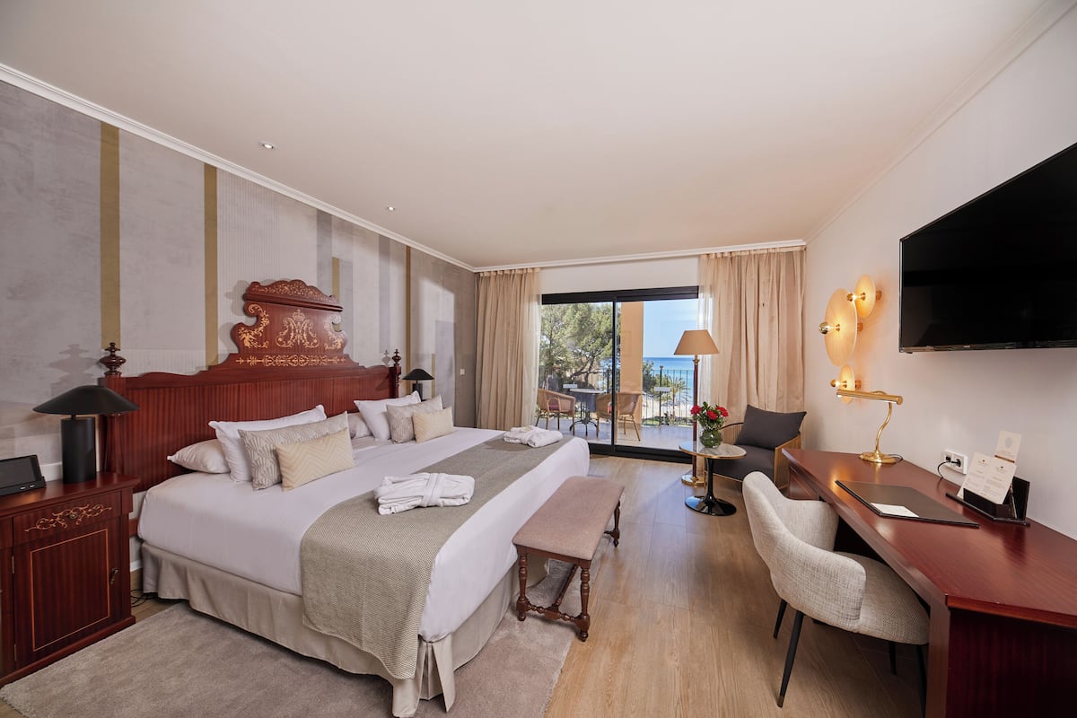 A hotel room with a large bed, a desk, a chair, a TV, and a balcony with outdoor seating.