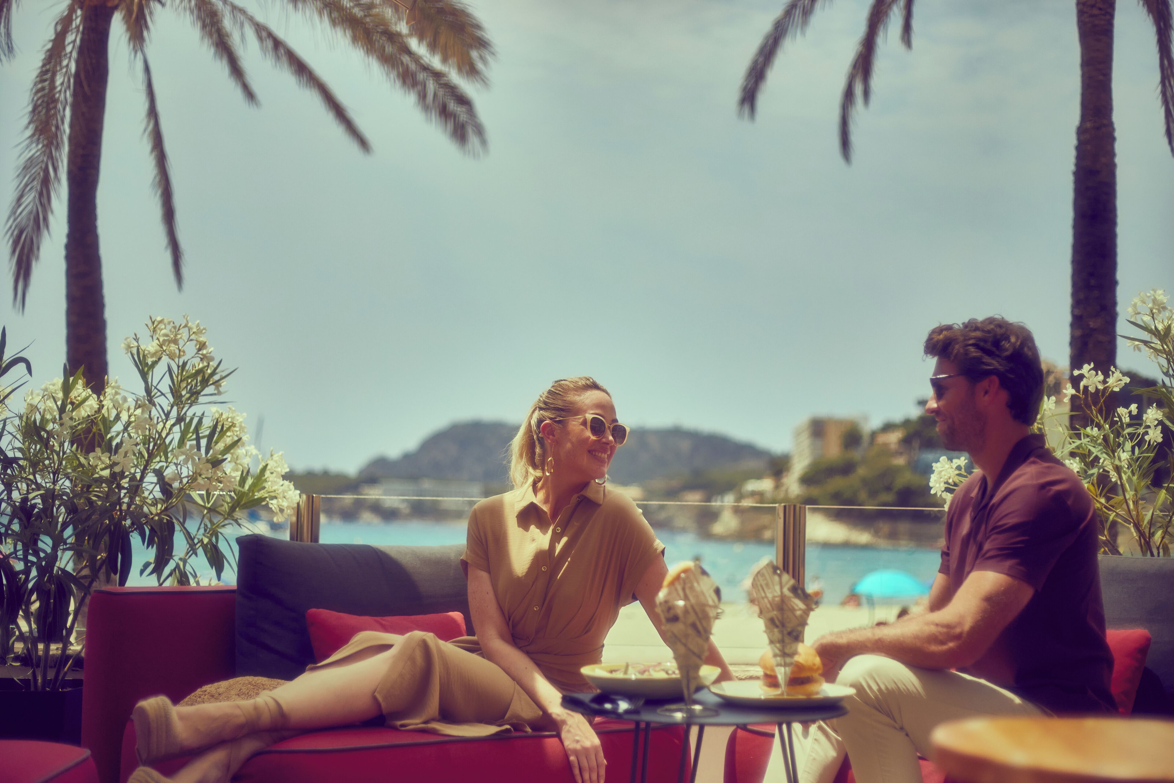 Una pareja sentada en un sofá rojo con una mesa entre ellos, con vista a una playa con palmeras y un cielo despejado.