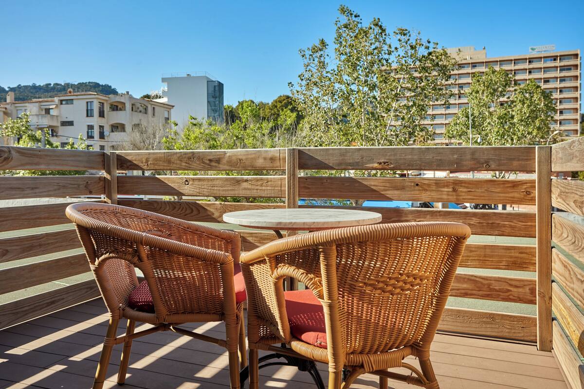 A wooden deck with wicker chairs and a round table, overlooking a pool and buildings.