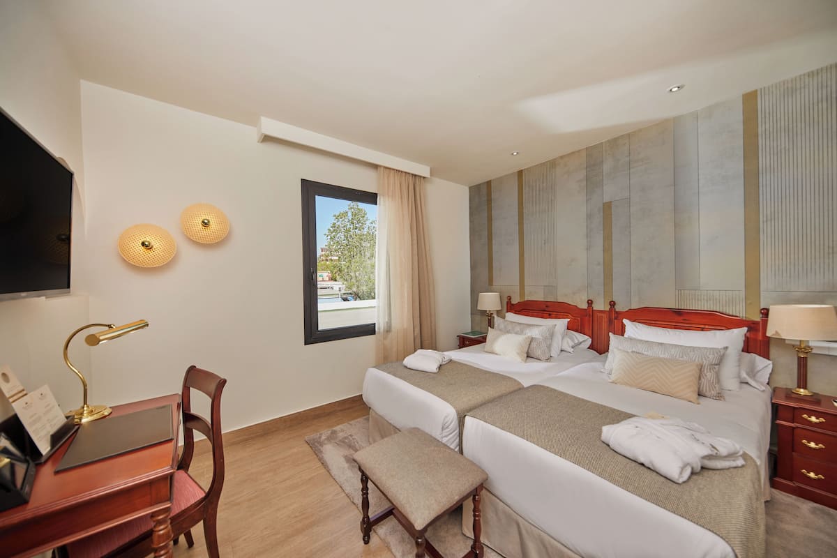 A hotel room with two beds, a desk with a chair, a TV, and a window with a view of the outside.