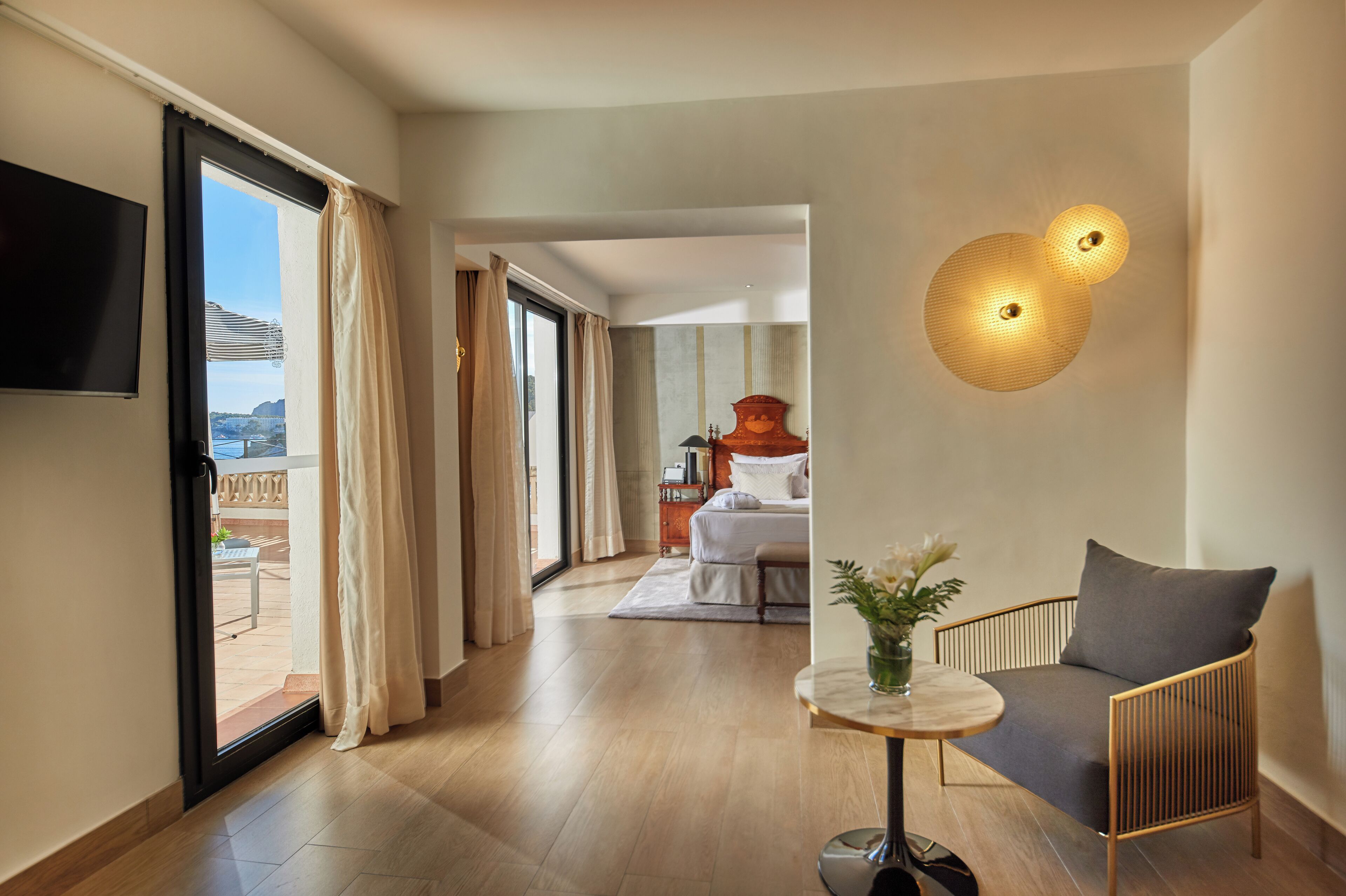 A hotel room with a bed, a TV, a table with a vase, and a chair. There is a view of the outside through the window.