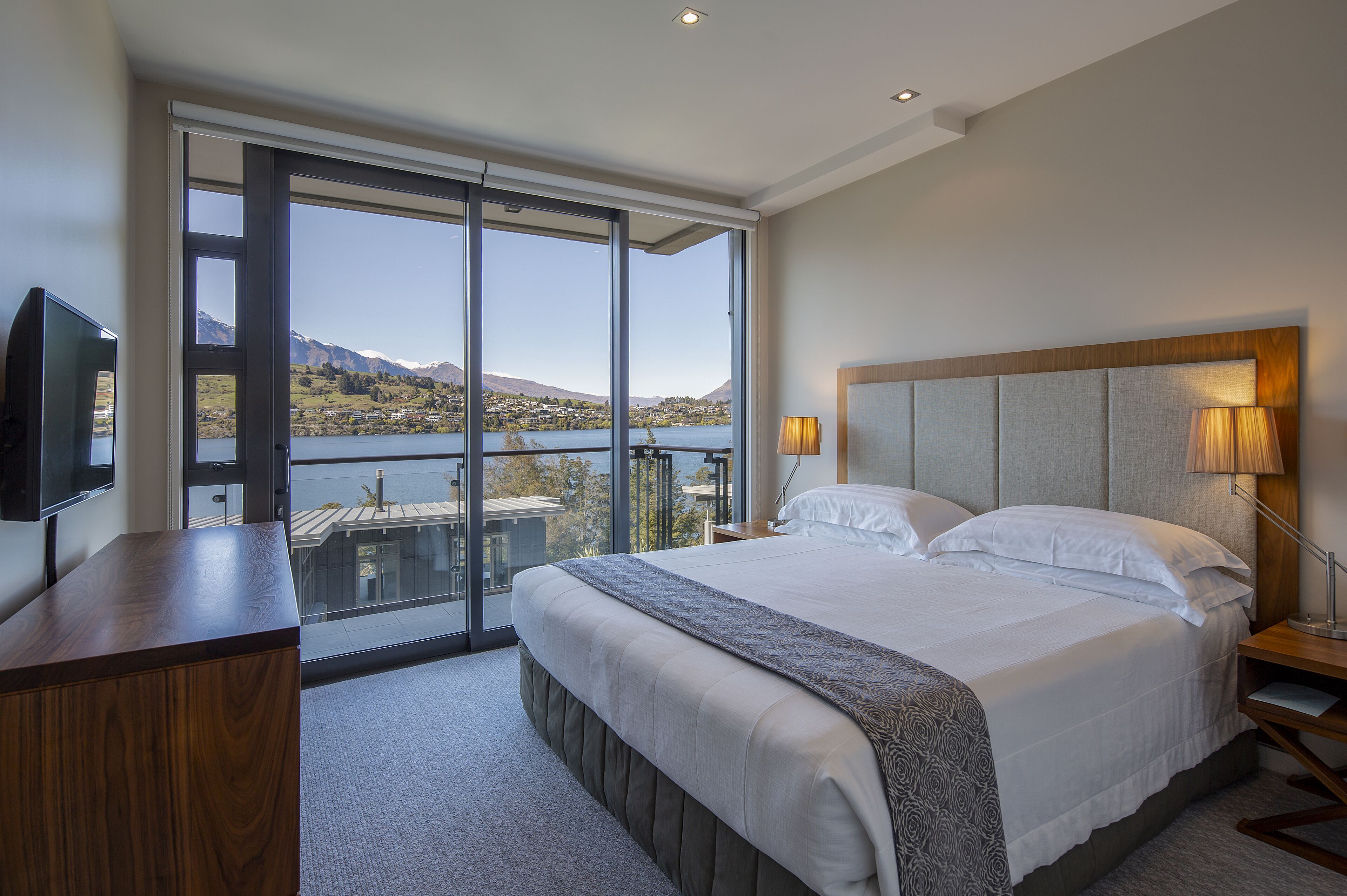 A bedroom with a large bed, a desk, and a TV. It has a view of a lake and mountains.