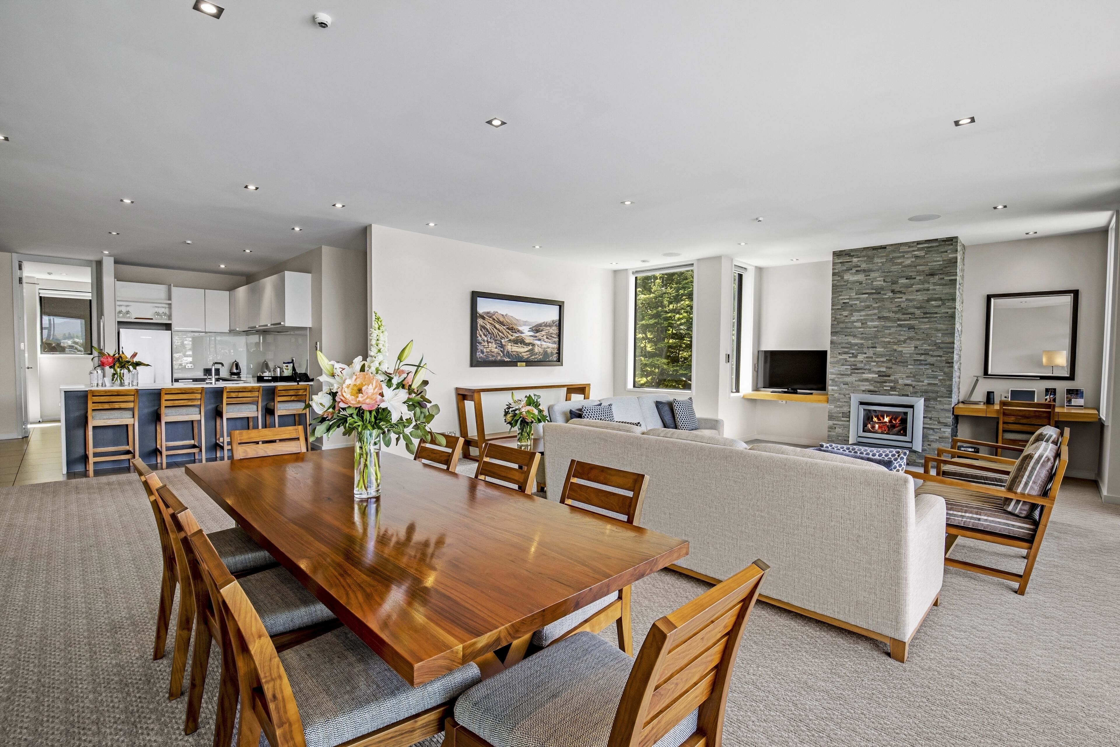 A spacious living area with a wooden dining table, comfortable seating, a fireplace, and a kitchen in the background.